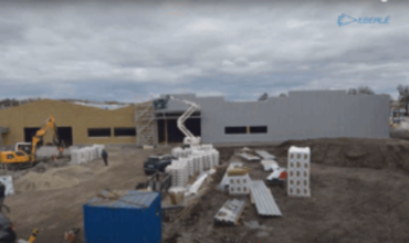 Chantier d’extension du site industriel d’Eberlé à Reichstett, avec engins de construction, échafaudages, blocs de béton et bâtiments en cours de finition sous un ciel nuageux.        