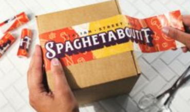 Mains appliquant un ruban adhésif imprimé « Spaghetaboutit » sur une boîte en carton, avec des ciseaux posés à côté.
