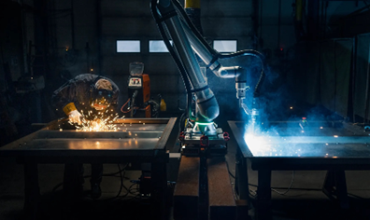 A Universal Robots collaborative robot performs an automated welding operation alongside a technician in an industrial workshop, illustrating human-machine collaboration and the precision of automation.