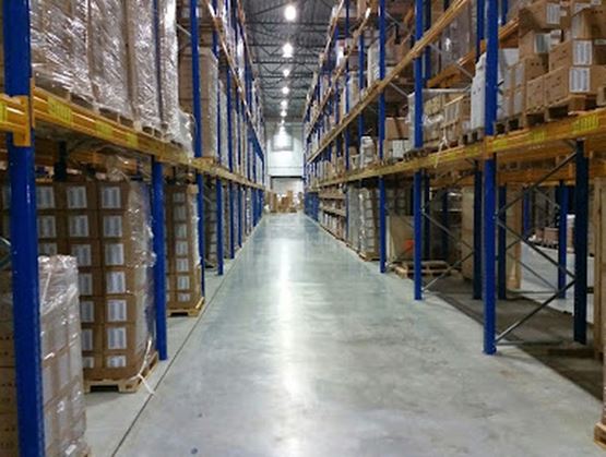 Central aisle of a logistics warehouse with blue and yellow metal shelving units loaded with pallets and boxes, under industrial lighting.