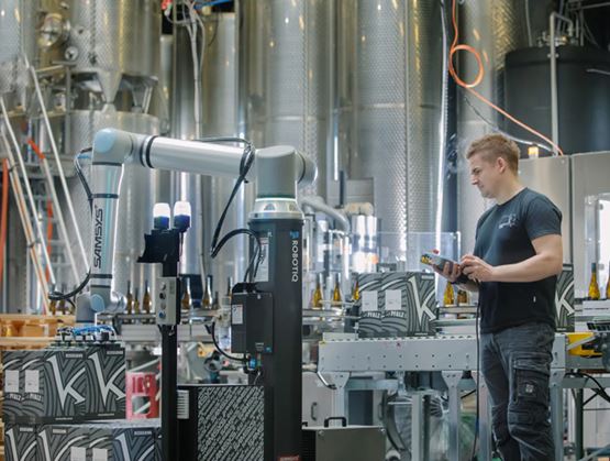 Technician operating a collaborative robot on an automated packaging line inside an industrial facility with large production tanks.