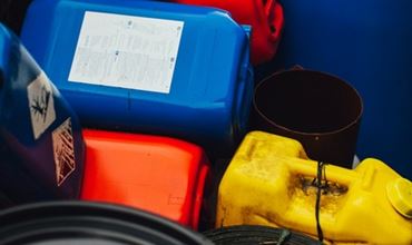 Several plastic jerrycans and containers in different colors (blue, red, yellow, and green) stacked outdoors, with a large blue barrel and rolled materials nearby, suggesting storage or industrial waste.