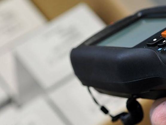 Hand holding a portable data entry terminal in front of labeled packages in the background.
