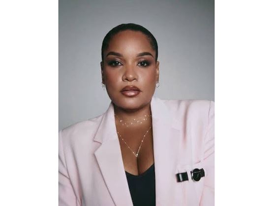 Portrait of a woman wearing a light pink blazer, a black top, and jewelry against a gray background.