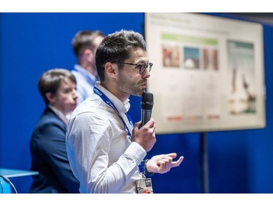 Man holding a microphone and speaking at a conference, with other participants and a presentation screen in the background.