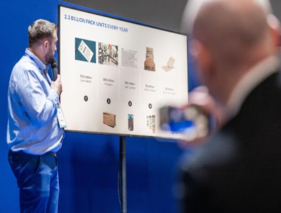 Professional presentation on packaging volumes, with a speaker holding a microphone in front of a screen and an attendee filming the conference.