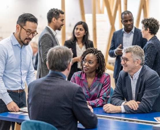 Cette image montre un groupe de professionnels qui discutent dans un espace de bureau moderne. Certains sont debout, tandis que d'autres sont assis autour d'une table
