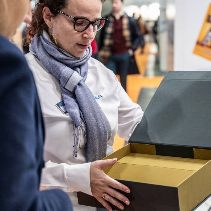 An exhibitor presents a premium rigid packaging box to a visitor during a trade show.